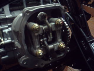 キャプチャー１２５のシリンダヘッドには他のＧＹ６エンジンには無い逆転デコンプ機構が装着されています。　これはエンジン停止時に圧縮上死点手前で僅かにカムシャフト（エンジン回転）が逆転することを利用し、ストッパーとワンウェイクラッチで構成した機構が僅かにデコンプカムを回転させてＥＸロッカーアーム（排気バルブ）を僅かに開け、始動時の最初の圧縮圧力を低減するのものです。　ＳＲやエンフィールドが最初の一蹴りの前に手動デコンプで圧縮抜く感じです。